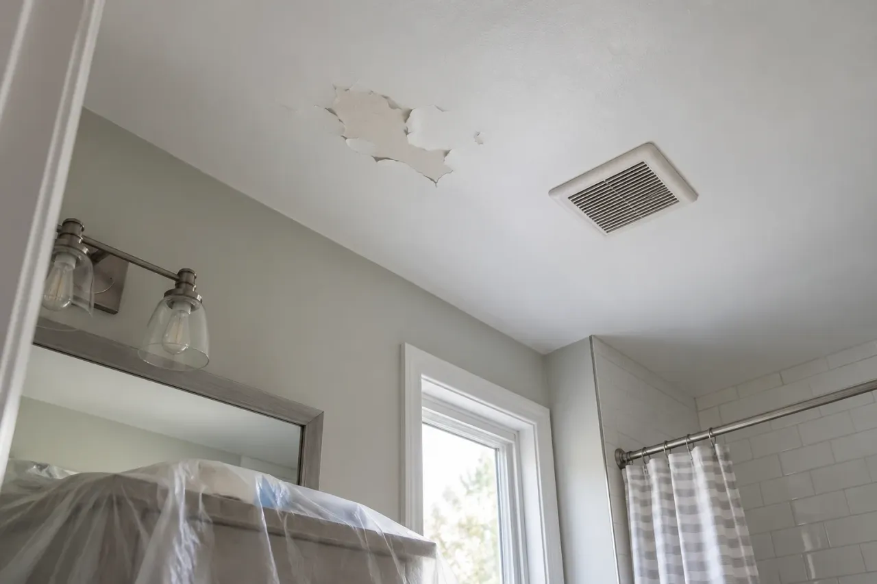 Peeling paint on a flat bathroom ceiling near an exhaust grille above a protected vanity